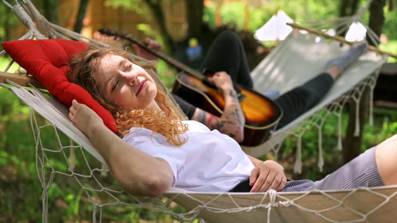 Couple lying on hammocks. Young man is playing guitar while woman is lying with closed eyes and smile. Greenery around. Glamping. Slow motion