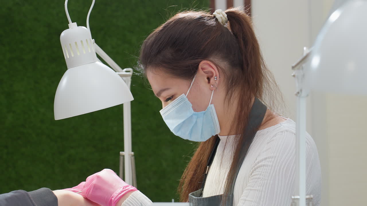 Closeup of nail technician in pink gloves filing customer nail while seated beside bright green wall under white desk lamp, focusing on precise grooming in calm salon environment