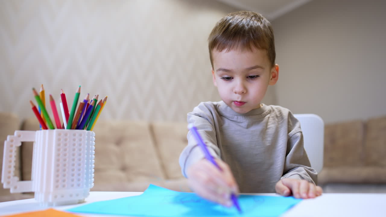 Child making art indoors. Young boy focused on drawing with colorful pencils on blue paper in a cozy room during the day