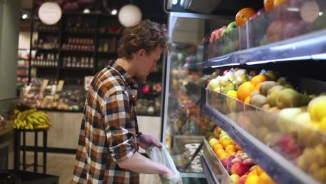 hombre seleccionando frutas frescas en el departamento de productos de la tienda de comestibles y poniéndolas en una bolsa de plástico. joven está eligiendo naranjas en el supermercado y poniéndolas en la cesta de la tienda. vista lateral