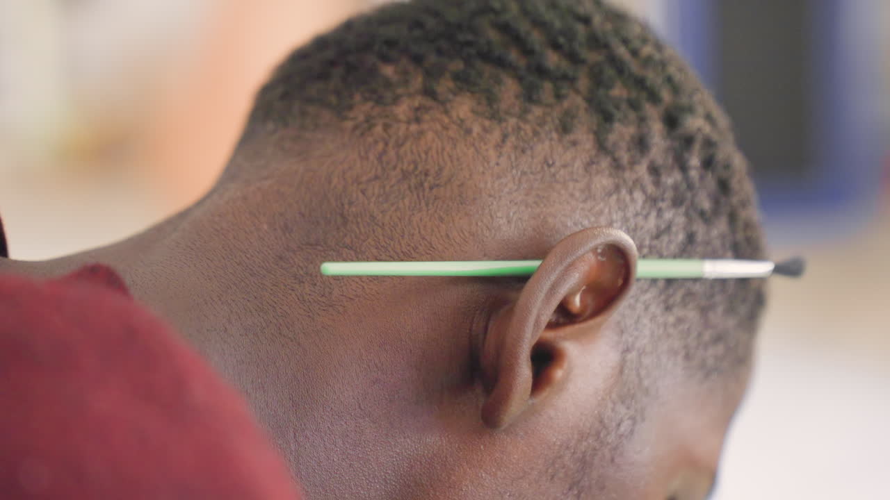 Studente artista di colore con un pennello verde dietro l'orecchio, chinato in avanti in un primo piano di profilo in classe, che mostra capelli corti e texture e la pelle, concentrato a dipingere la tela con un'espressione attenta.