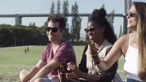 smiling friends clinking beer bottles while sitting on meadow.