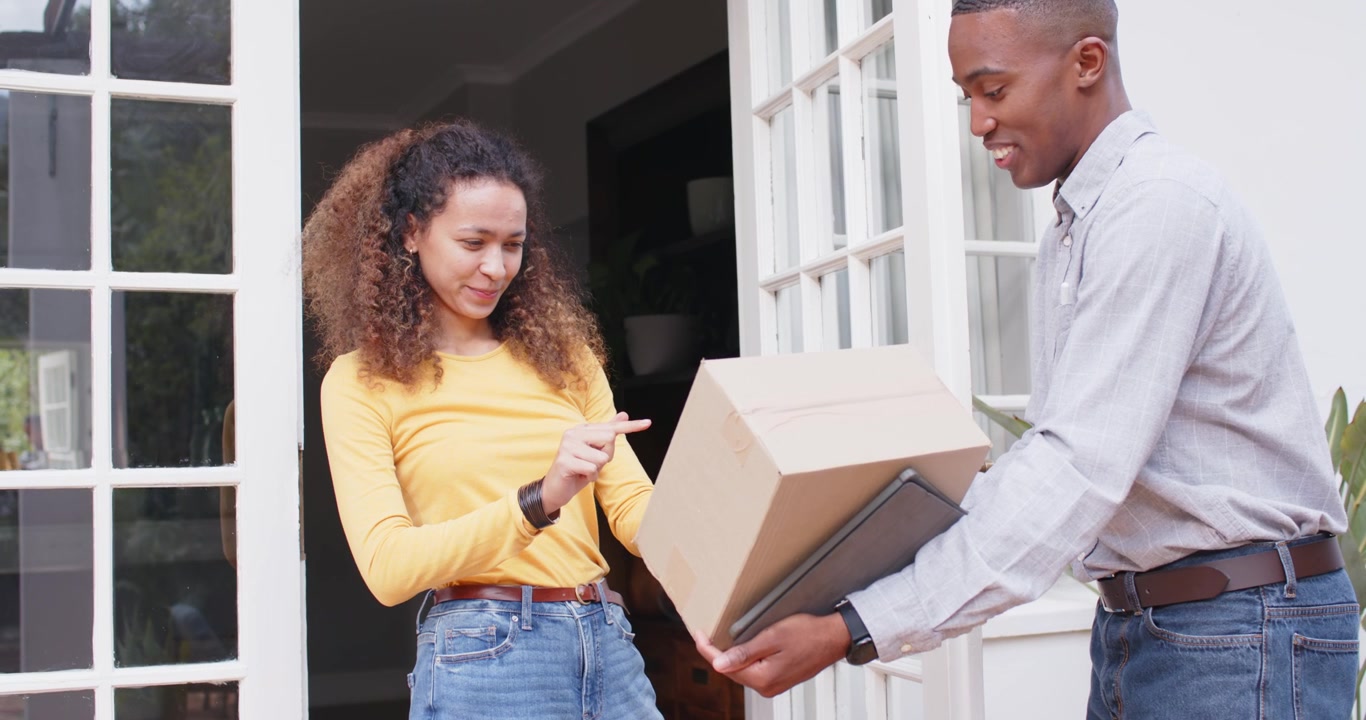Diverse courier and woman at porch holding box and tablet, woman tapping phone confirming delivery