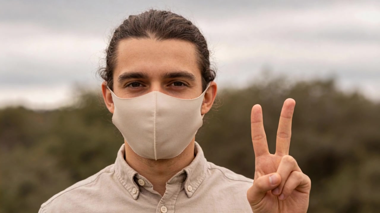 Hombre levantando la mano derecha, con mascarilla facial beige y camisa beige, haciendo el signo de la victoria, posando en el parque.