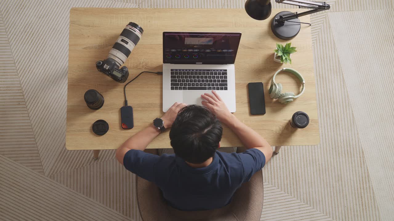 Top View Of A Male Color Grading Shaking His Head And Having A Headache While Sitting In The Workspace Using A Laptop Next To The Camera Editing The Video At Home