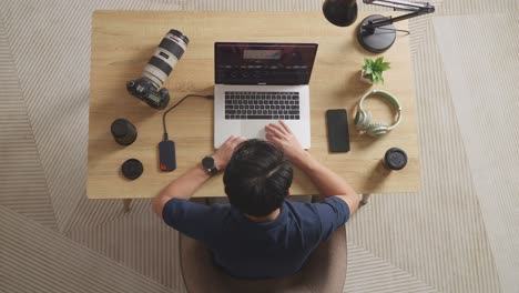 top view of a male color grading shaking his head and having a headache while sitting in the workspace using a laptop next to the camera editing the video at home