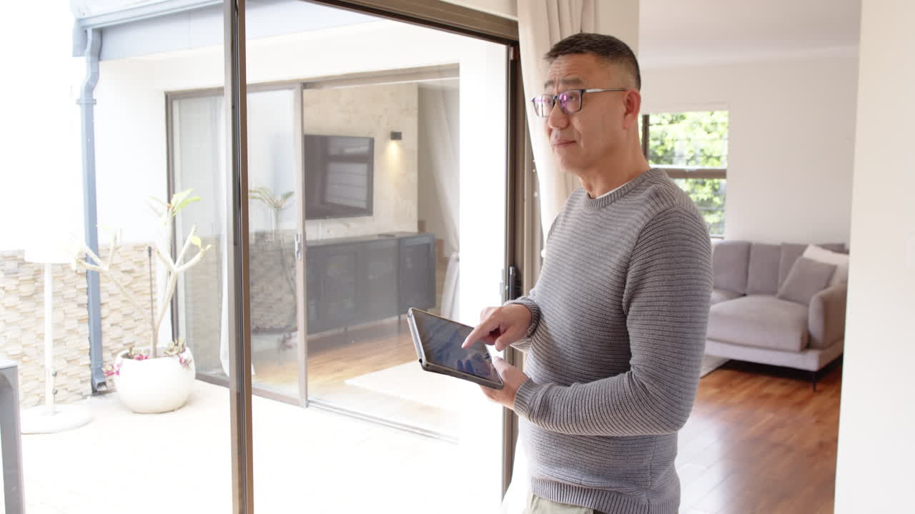 Man using tablet at home, standing by large window, wearing glasses, copy space