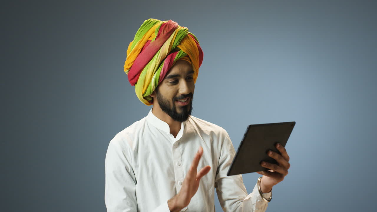 Young cheerful Indian man in traditional clothes and turban making a video call using a tablet