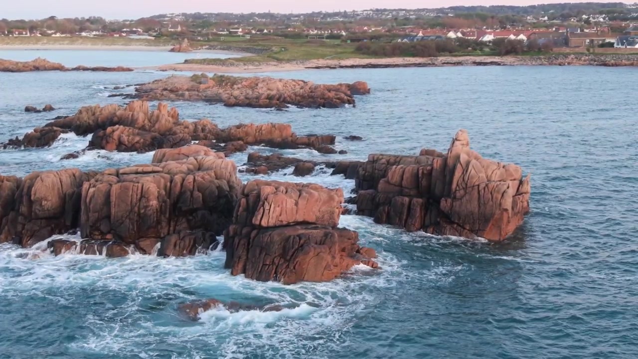 Guernsey, Isole del Canale, riprese ravvicinate a bassa quota con drone di una costa frastagliata sopra onde spumeggianti che si infrangono su affioramenti rocciosi di granito sotto il sole del tardo pomeriggio con il rosa che inizia a tingere l'orizzonte