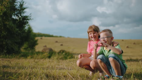 siblings squatting outdoors in bright sunlight as sister holds brother close, both smiling joyfully while mum captures their playful moment with a photograph, surrounded by open countryside and lush