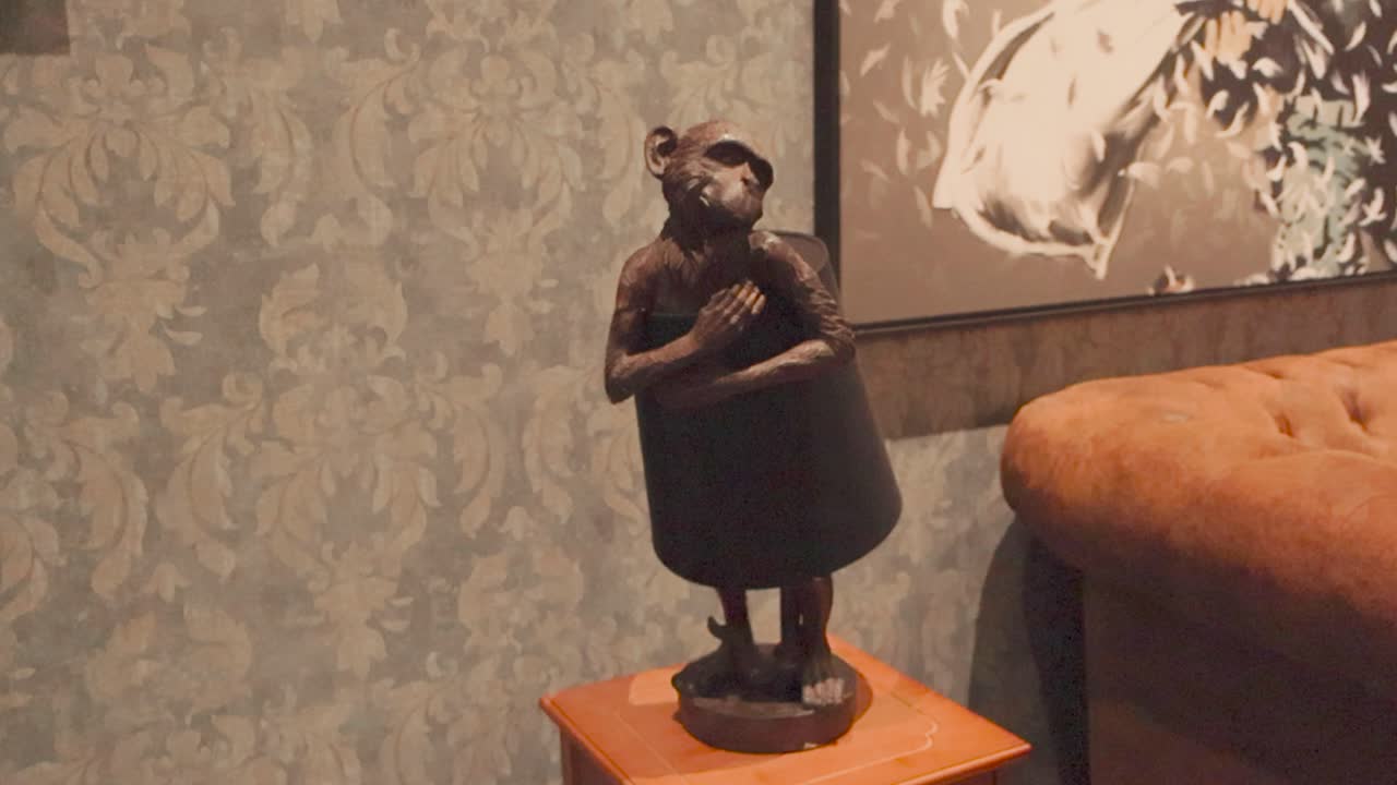 Footage shows a vintage and metallic dark monkey lamp or a lampshade standing on a small table, while an orange sofa and vintage wallpaper is in the background at Banksy art exhibition.