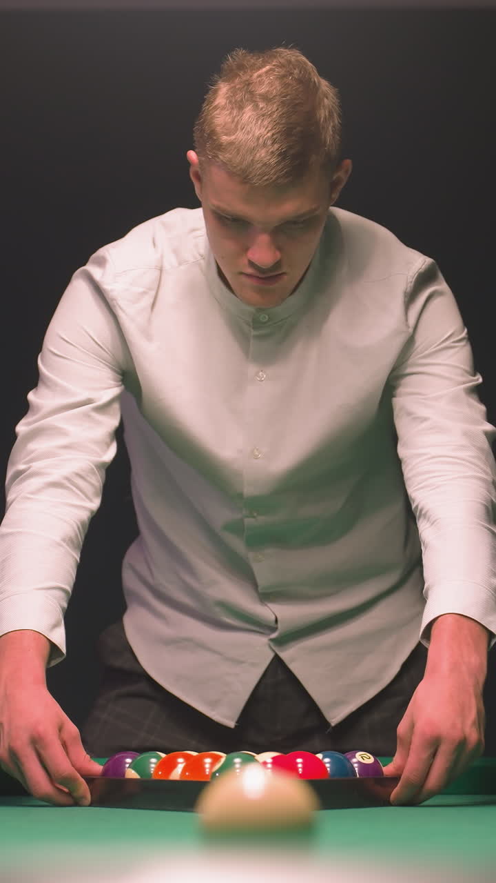 primer plano de un aprendiz de billar en camisa intensamente enfocado en la mesa de billar mientras se prepara para romper. sus manos se mueven sobre un estante de bolas coloridas en fieltro verde vibrante, luz suave contra un fondo oscuro
