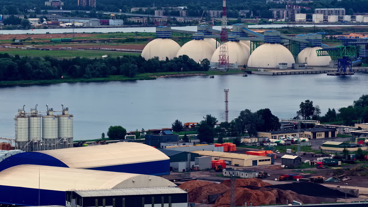 European industrial port with storage domes, silos, and waterway near forested urban edge in Riga, latvia