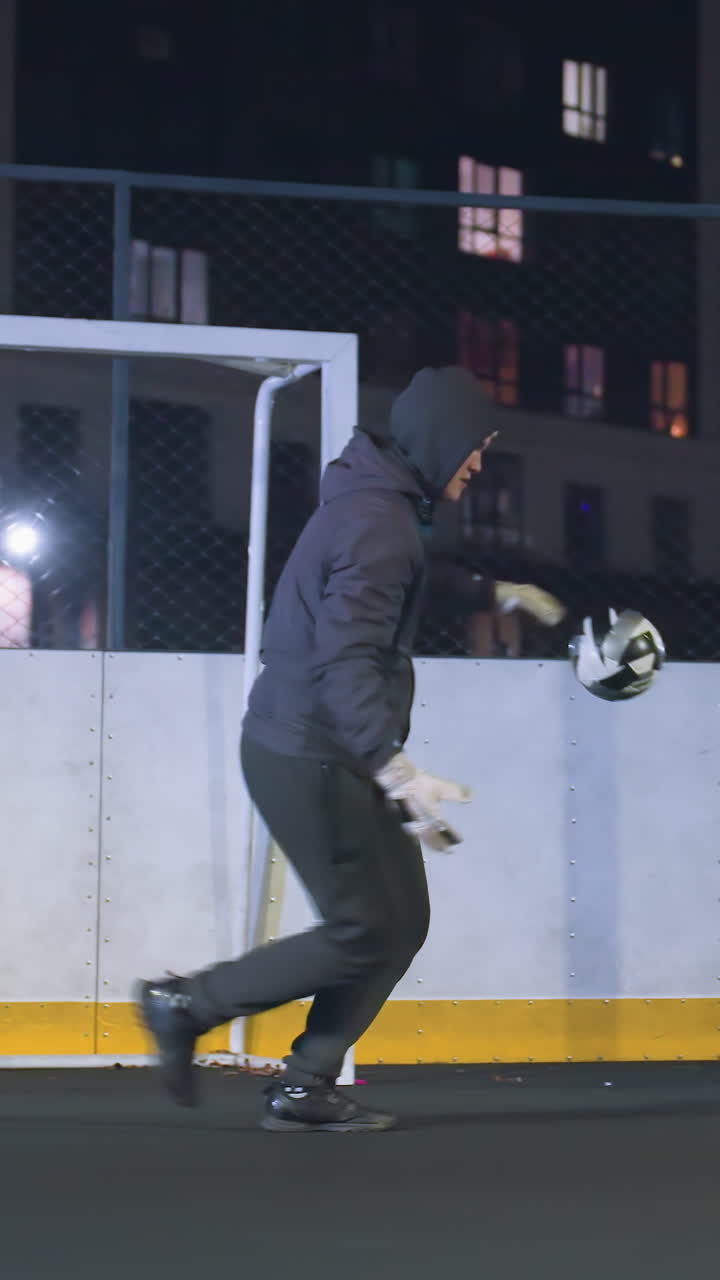 Goal keeper in a hoodie kicks soccer ball during night practice on an urban outdoor field, surrounded by tall buildings, the action highlights athletic skills under bright floodlights