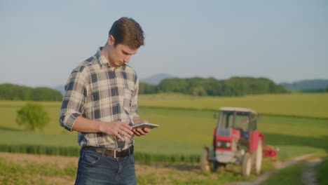 trendy farmer works on digital tablet near tractor and fields