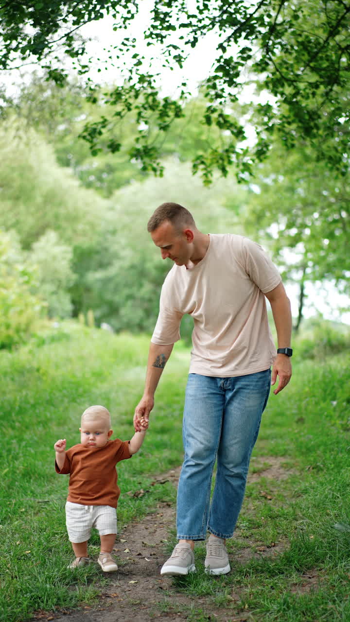 Father holds his little son by the hand walking by the park. Teaching a child to walk outdoors. Vertical video.