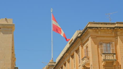 the flag of the knights of st john flies proudly above the buildings of malta - its red and white cross a strong contrast to the blue sky above