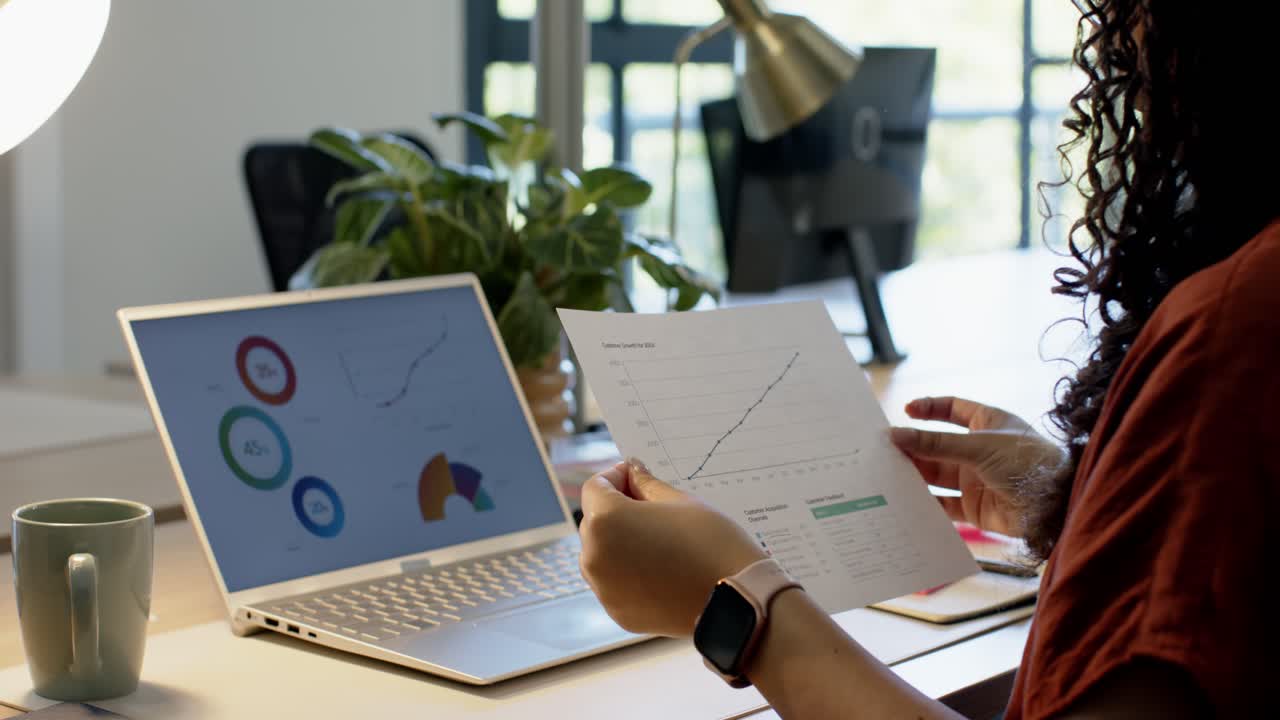 Adjusting glasses, woman is comparing paper charts to laptop screen while checking trends