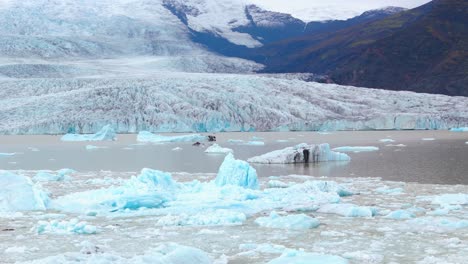 Iceberg-Azul-Turquesa-Derritiéndose,-Al-Fondo-El-Glaciar-Y-Sus-Montañas-De-Islandia
