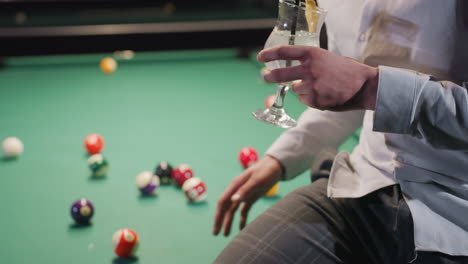 person in white shirt and grey trousers holding lemon drink with black straw, sitting on green pool table with colorful billiard balls. casual setting, stylish attire, dim ambiance, and relaxed mood