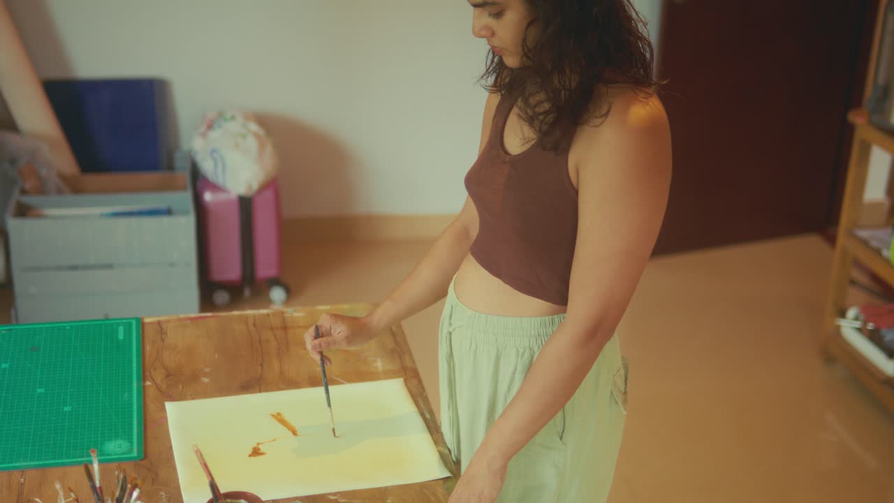Mulher pintando em papel com pincel à mesa em um estúdio aconchegante; cena interna de estilo de vida criativo.
