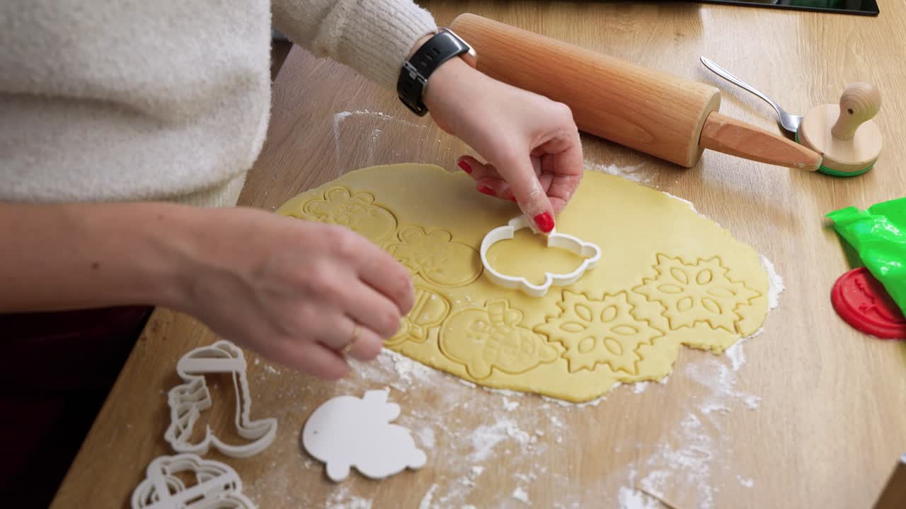 A close-up shot of hands pressing cookie cutters into dough, creating festive shapes. The scene captures warm natural lighting on a wooden table with baking tools nearby