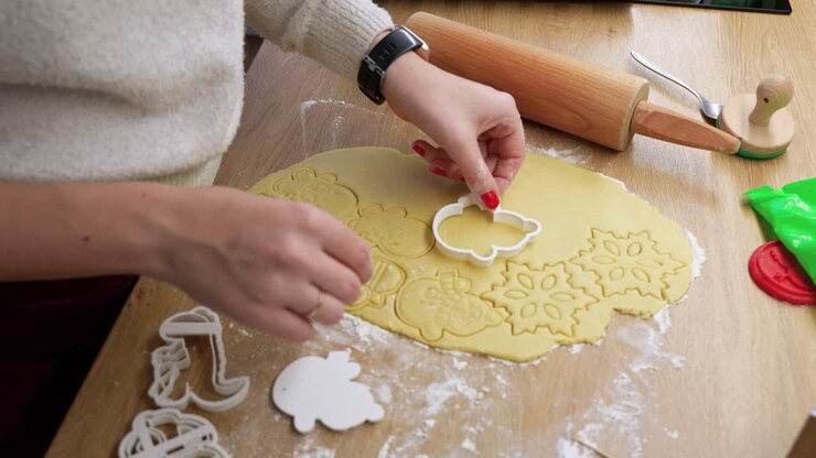 A close-up shot of hands pressing cookie cutters into dough, creating festive shapes. The scene captures warm natural lighting on a wooden table with baking tools nearby