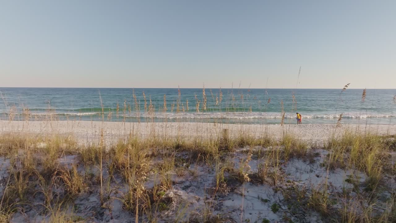 Daisy cutter pan drone shot of a couple walking on an empty white sand beach along the sea, with weeds in the foreground and clear blue sky in the background, creating a serene and peaceful scene
