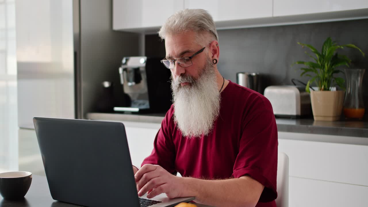A serious and confident elderly man with gray hair and a lush beard wearing glasses and a red T-shirt is typing on a gray laptop while working remotely in a modern apartment