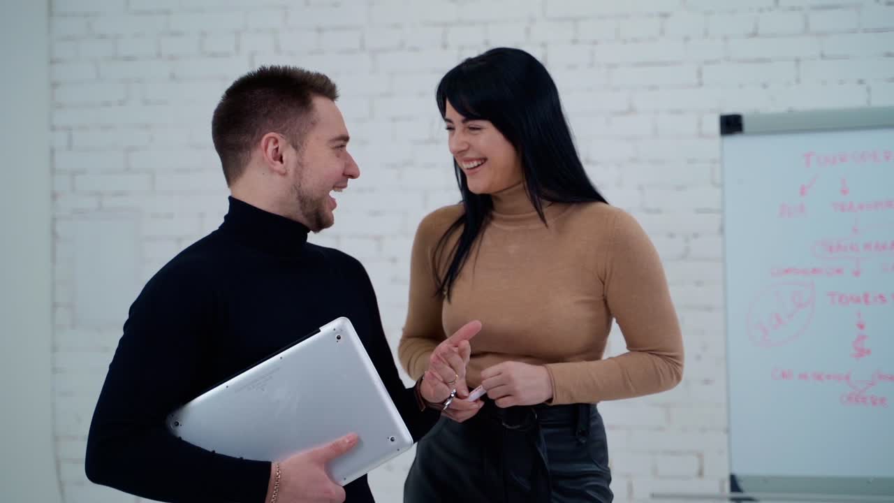 Smiling coworkers talking indoors. Handsome man holding a laptop and telling something to a young woman standing near the board for writing.