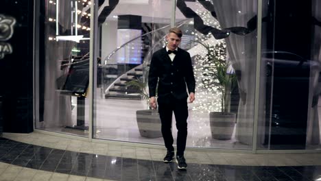 attractive young man in a black suit walking out of a hotel in the evening. behind a lot of blurred lights and glass door of luxurious hotel