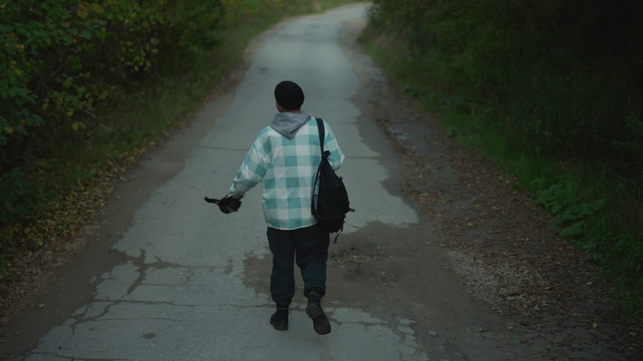 white male hiker walking on road, carrying camera and tripod, backpack slung over shoulder, cracked pavement and damp patches, overcast sky, quiet rural lane, slow reflective pace, autumn leaves