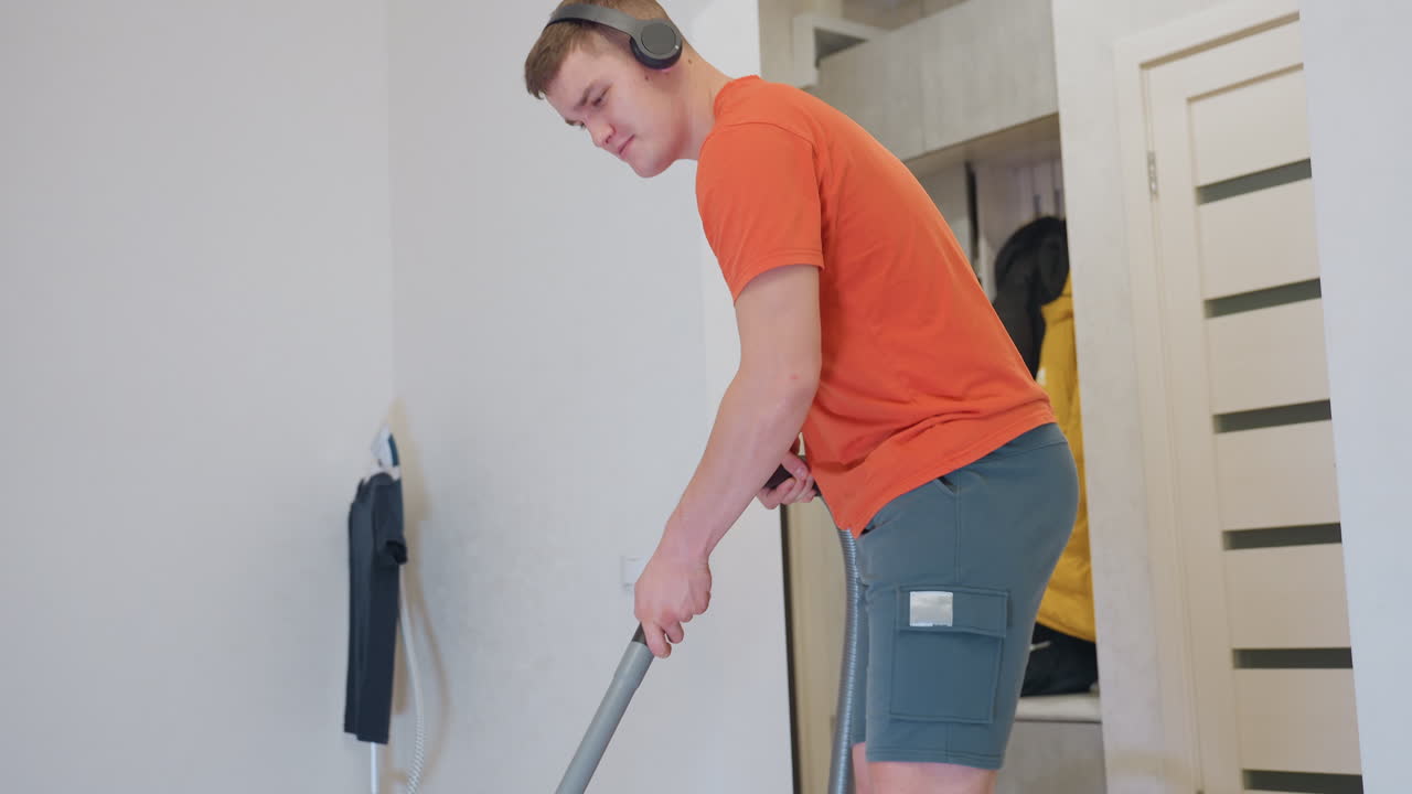 Young man in orange shirt and shorts enjoys music while vacuuming floor, moving with rhythm and dancing during household chore, concept of multitasking