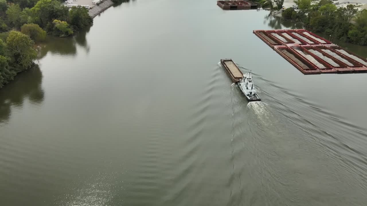 Aerial drone footage of towboat guiding cargo barge along calm Kanawha River surrounded by forested hills, industrial docks, and quiet roads in scenic West Virginia, United States