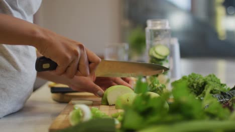 Mid-section-of-mixed-race-woman-preparing-healthy-drink,-cutting-fruit-and-vegetables-in-kitchen