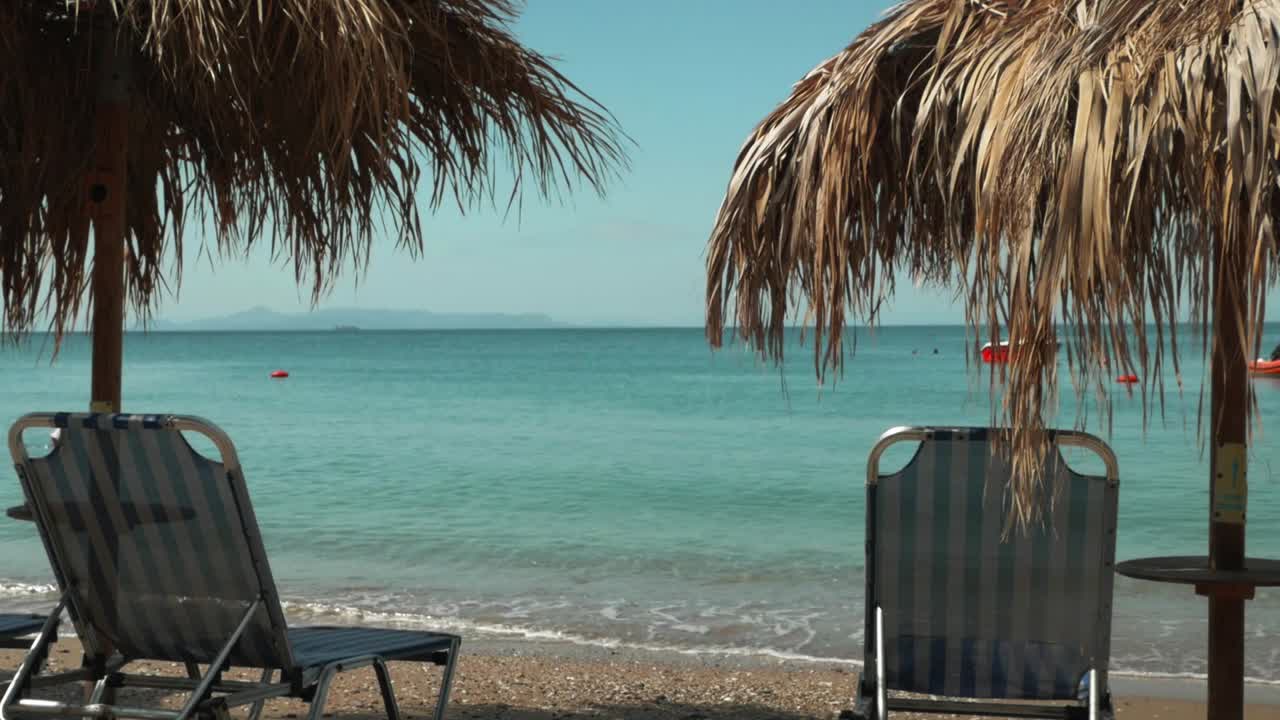 Spiaggia greca sabbiosa con lettini da spiaggia e ombrelloni di palma, mentre l'acqua cristallina e azzurra accarezza la costa.