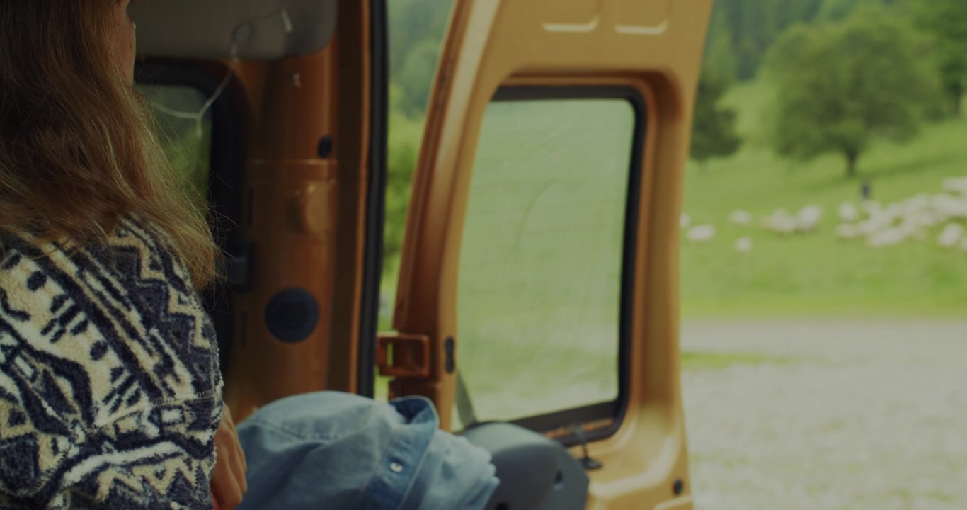 A closeup shot of a woman with long hair next to an open camper van door, looking out at a green mountain landscape with sheep grazing.