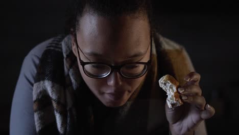 mujer negra comiendo un sándwich en la cocina oscura mientras se inclina sobre la mesa