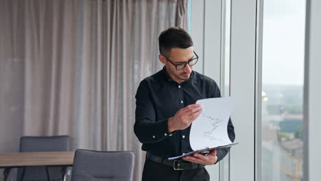 businessman in black shirt and glasses stands at the window looking through the documents. worried and annoyed man dissatisfied with data he sees in papers.