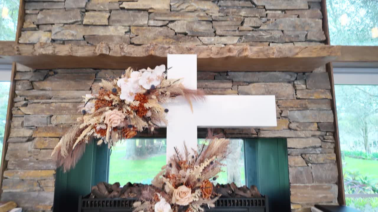 A wedding ceremony venue is decorated with white pillars, orange and white flowers, and a large, decorative cross