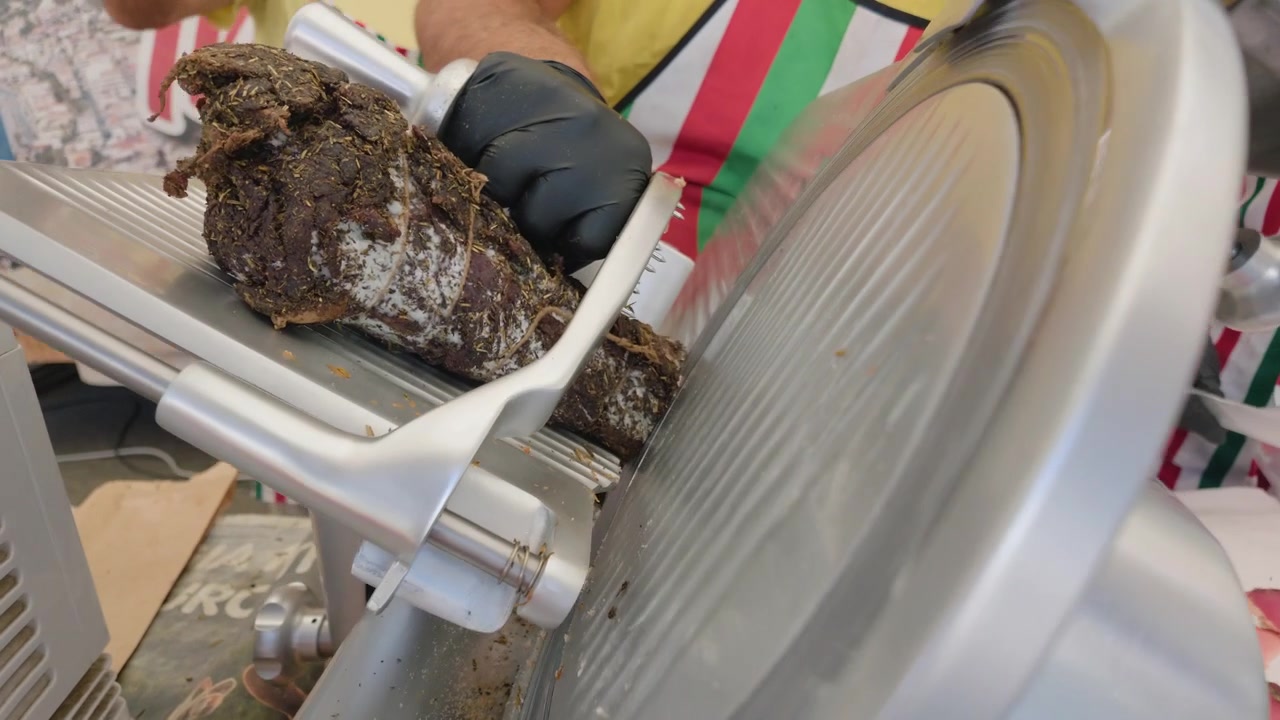 Primo piano di un salume alle erbe su un'affettatrice, con fette di carne su carta accanto.