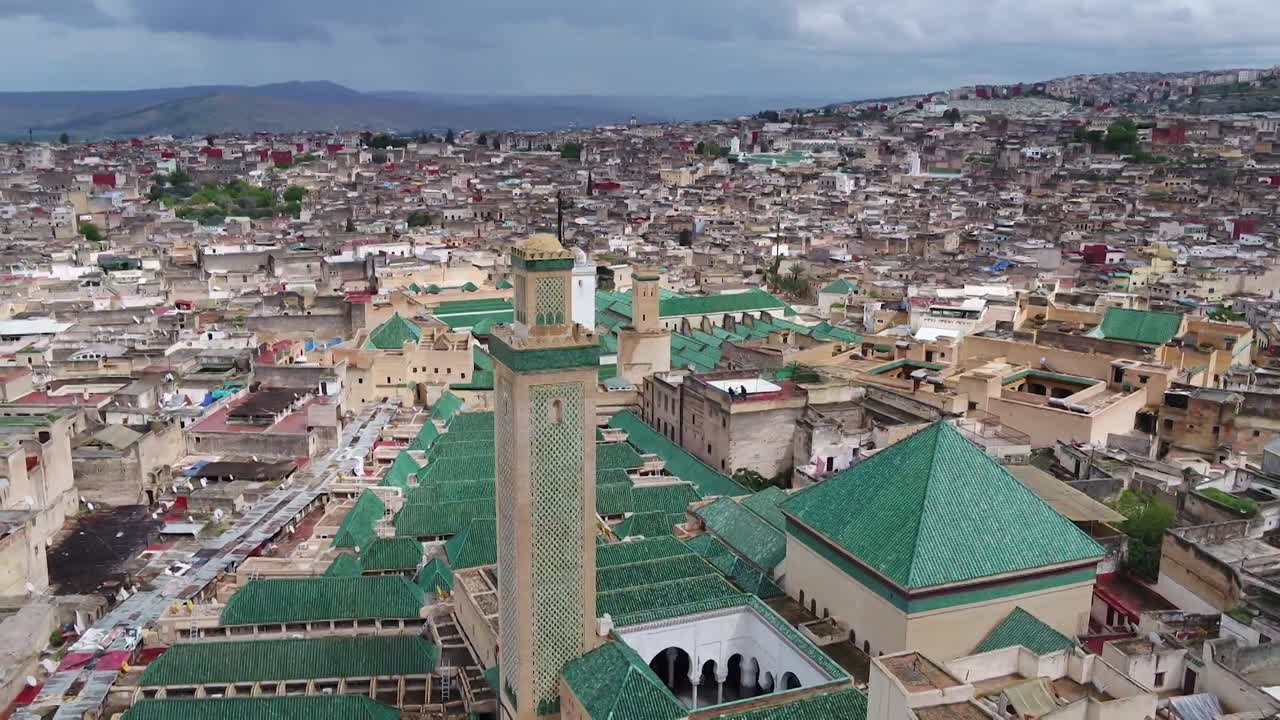 Exquisito primer plano de dron de un minarete de mezquita decorado con azulejos Zellige verdes. Presenta los icónicos tejados de tejas verdes y una vista panorámica de la histórica ciudad de Fez El Bali bajo un cielo nublado