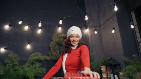 beautiful girl with christmas presents indoors. portrait of a happy young woman wearing red sweater, knitted cap and scarf throws gift on christmas background at home.