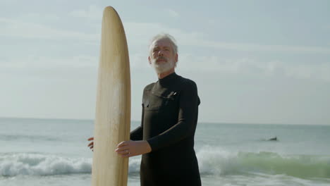 portrait of a senior man with surfboard standing on the sandy beach and looking at the camera