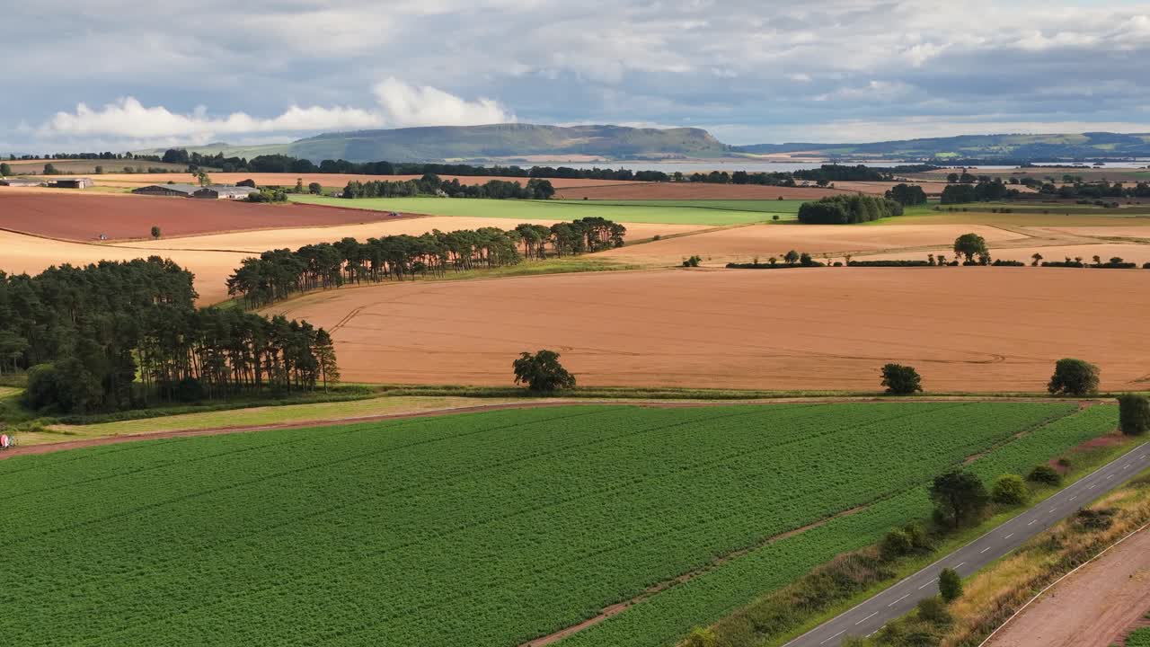Aerial camera glides over patchwork fields, green crops, and golden harvest under cloudy summer skies near Bennachie, Kinross, Scotland. Smooth lateral movement, natural daylight