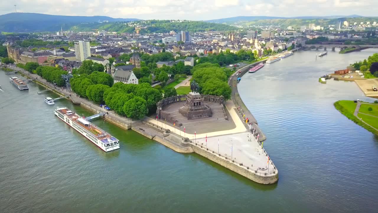 Aerial view of Deutsches Eck in Koblenz, Germany, where the Rhine and Moselle rivers merge. The historic monument, lush greenery, and passing cargo boats create a dynamic and scenic riverside scene.
