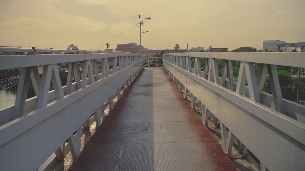 A steel pedestrian bridge stretches over a Bangkok canal during golden hour