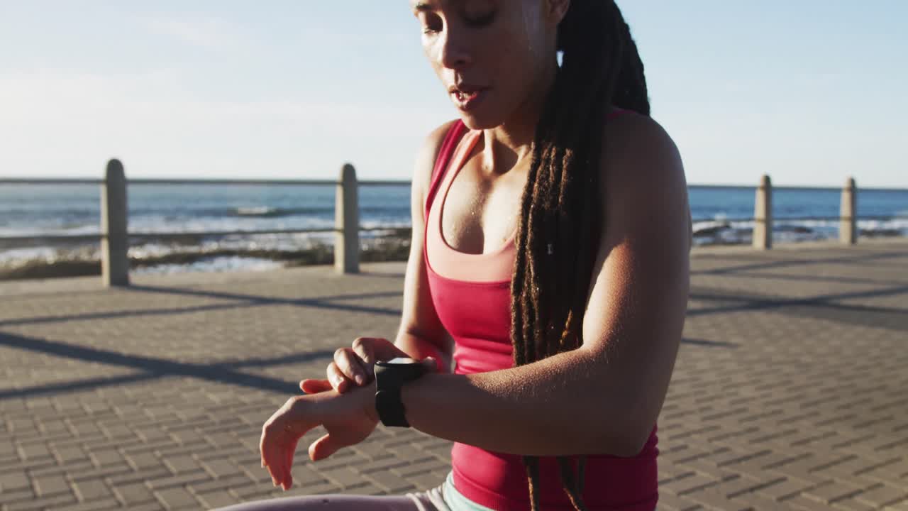 African american woman in sportswear checking smartwatch on promenade by the sea
