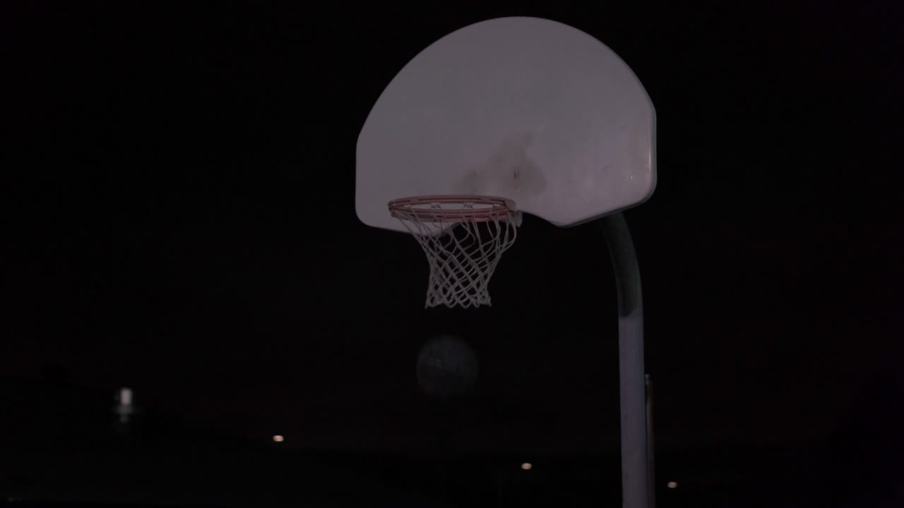 una adolescente regatea el baloncesto y luego lo tira en la red en una cancha al aire libre por la noche