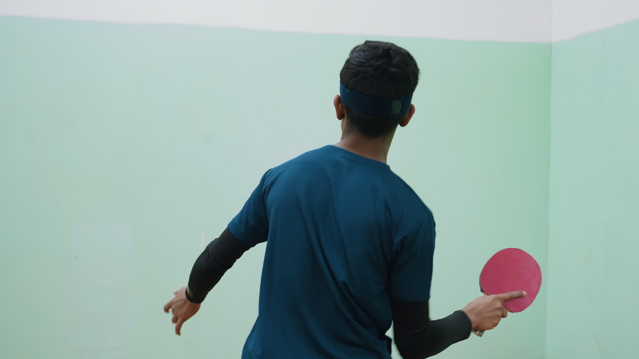 Back view of young athlete wearing headband and sportswear practicing tennis against wall indoors, holding paddle in active stance, training skills with determination during practice session